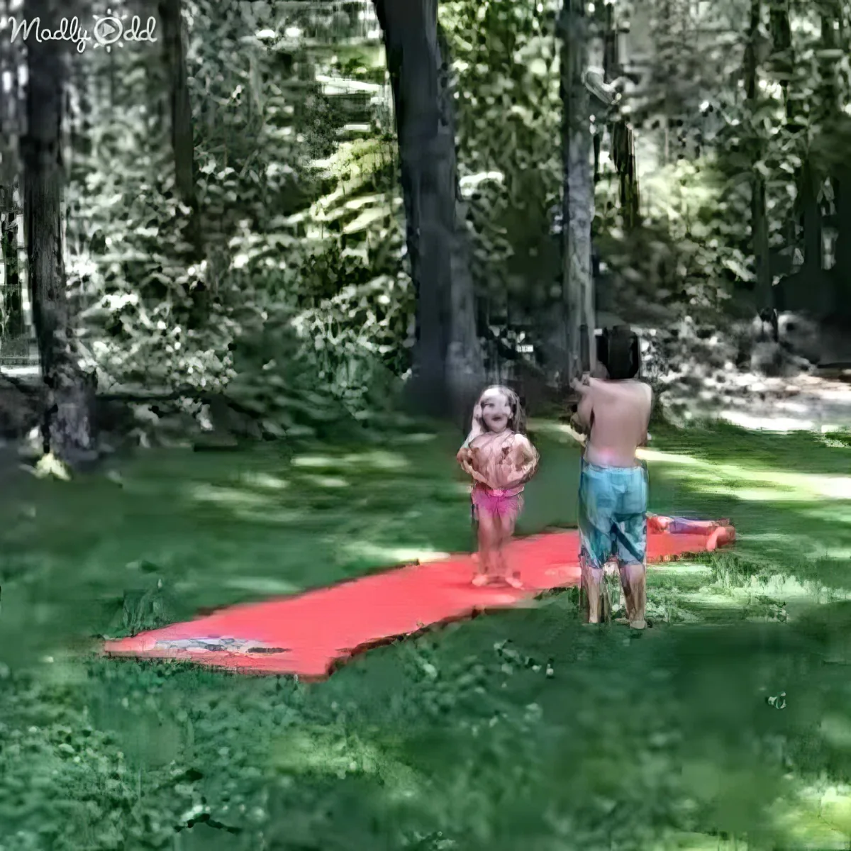 Two children stand on a red mat in a grassy area surrounded by trees.