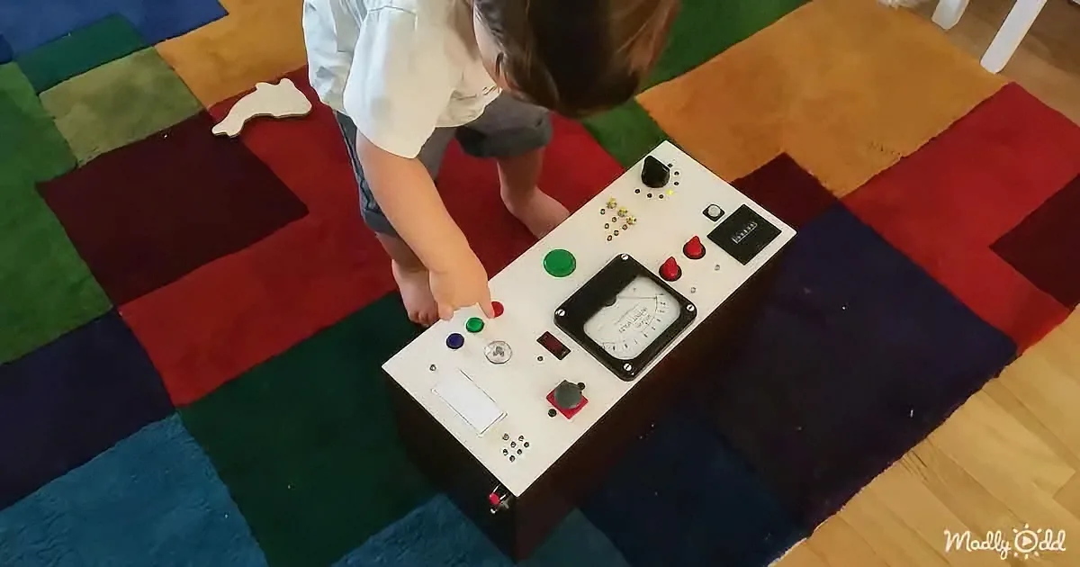 A child interacts with a control panel featuring various buttons and a gauge on a colorful rug.
