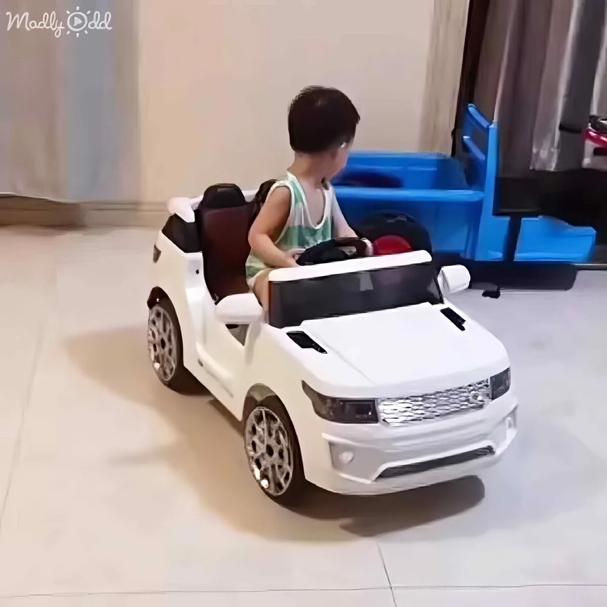 A child is driving a white toy car indoors, wearing a green tank top and shorts.