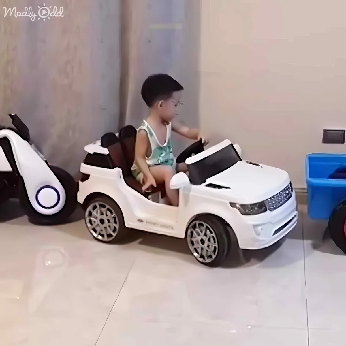 A child sits in a white toy car, playing indoors with other toys around.