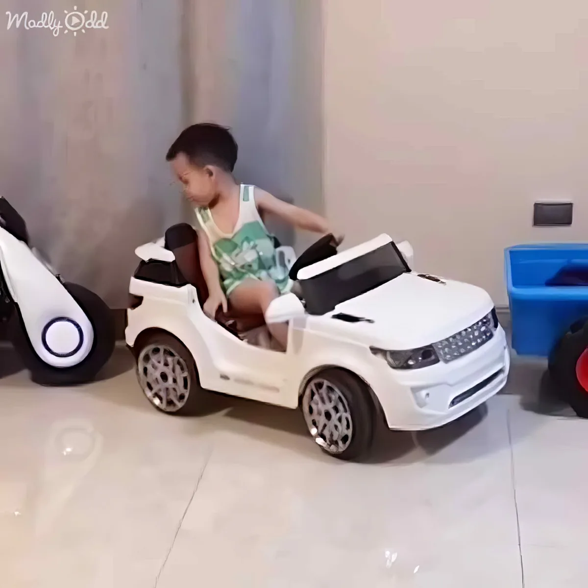 A child sits in a white toy car, appearing to struggle with the seatbelt while surrounded by other toy vehicles.