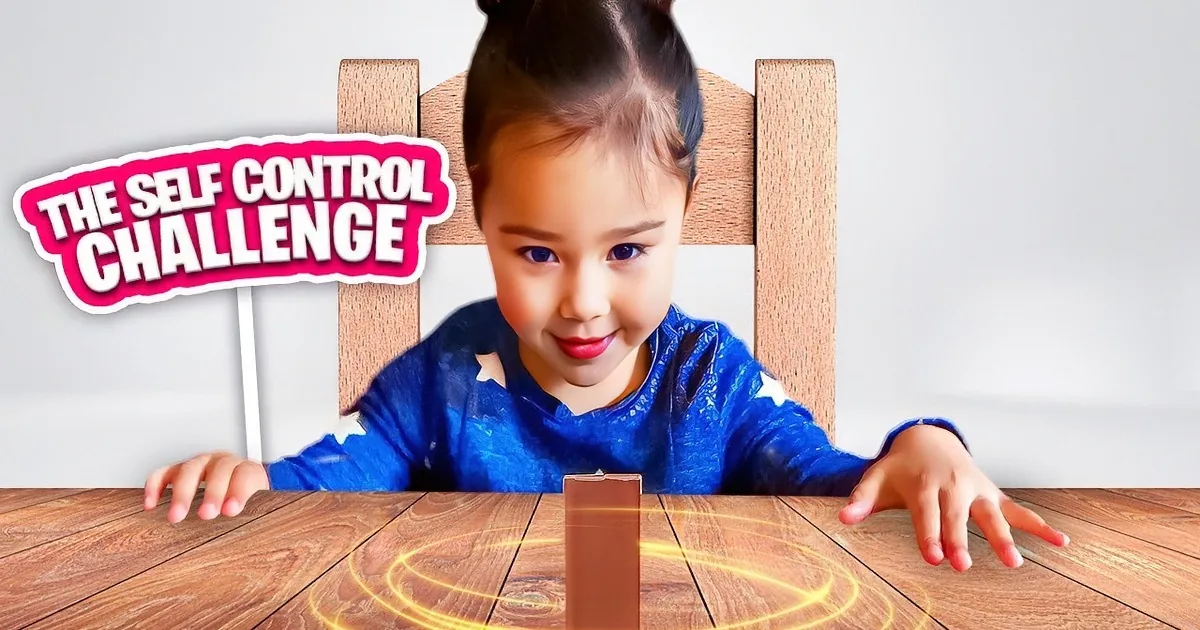 A young child with pigtails sits behind a table, smiling at a chocolate bar on a stand.