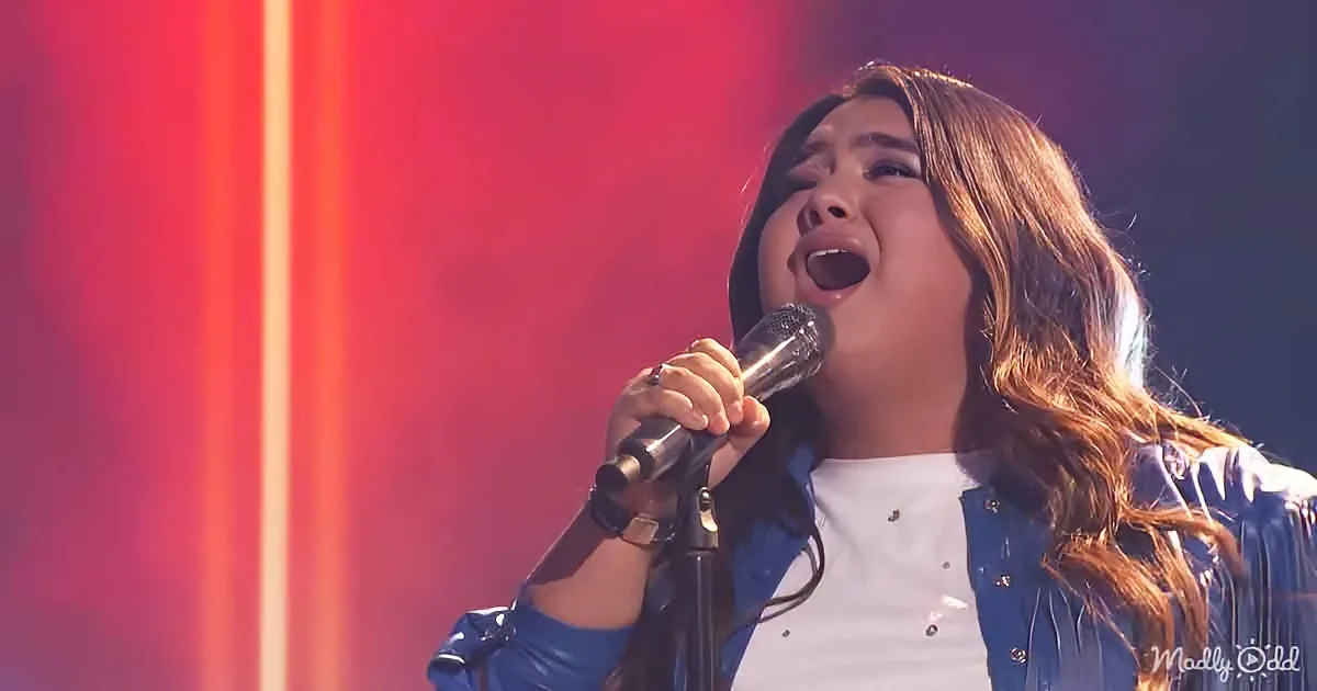 A person sings passionately into a microphone, wearing a blue jacket and white shirt, against a red and orange backdrop.