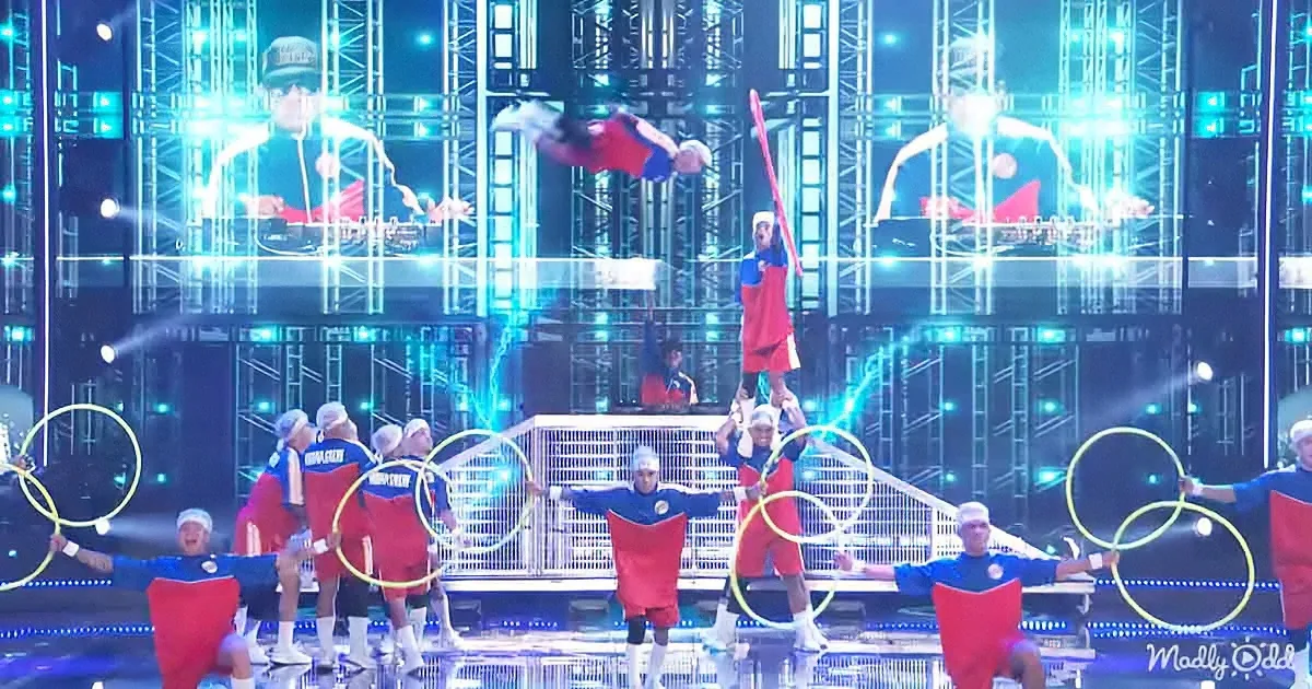 A performer in red and blue leaps mid-air, surrounded by athletes in similar attire and Olympic rings, under bright stage...