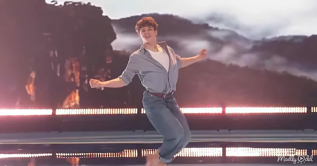 A person in a light blue shirt and jeans dances on a stage with a mountainous backdrop and red lights.