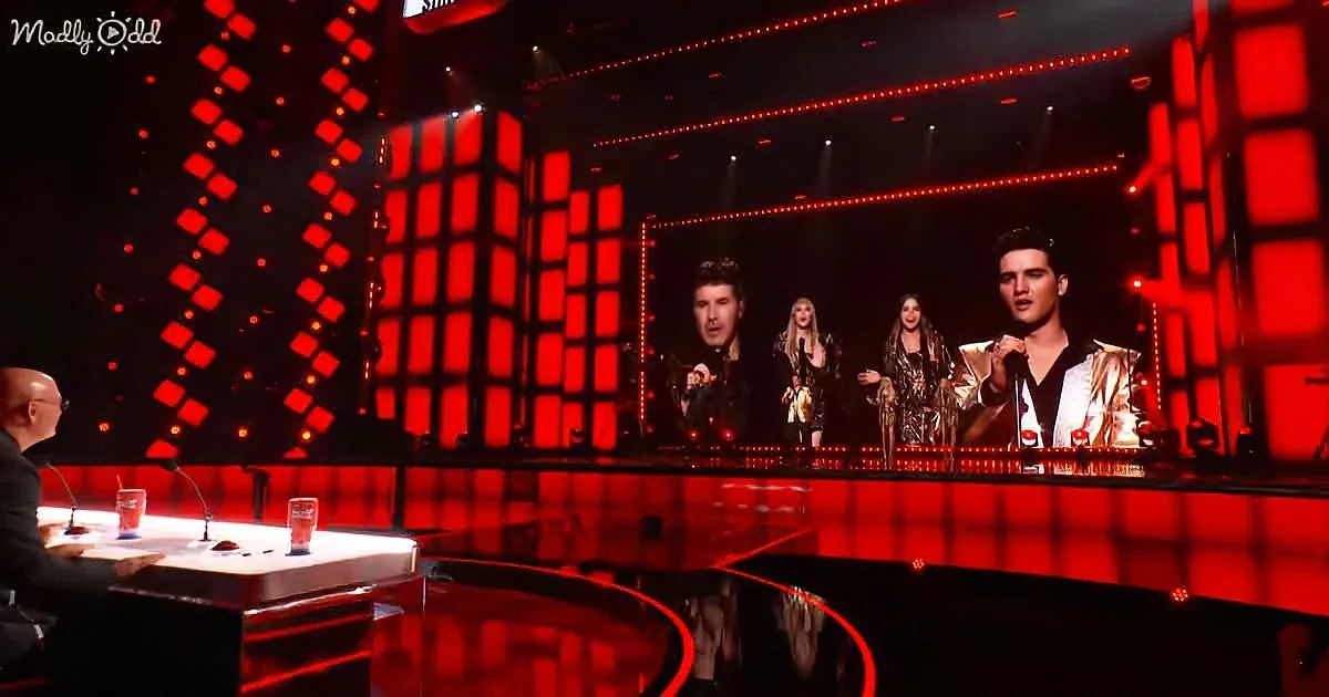 A performer in a gold suit sings on stage with judges and a red-lit backdrop.