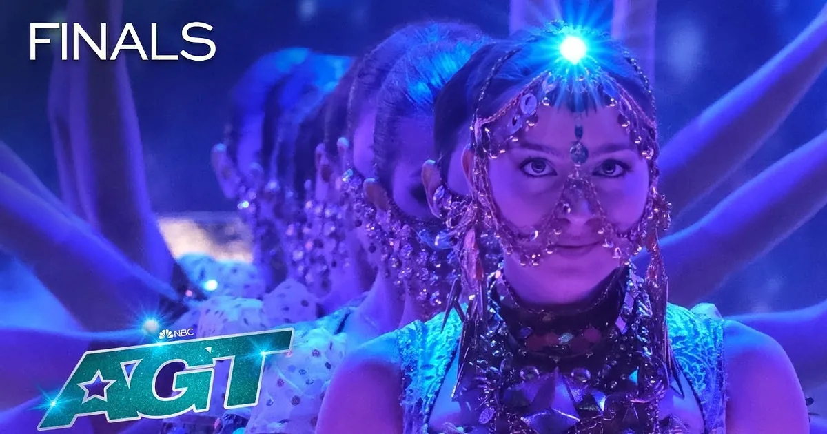 A dancer in a glowing, fluffy, galaxy-inspired dress stands on a stage with purple and blue lighting.
