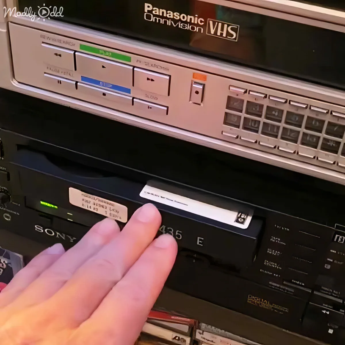 A hand reaches for a Sony VHS tape labeled "435 E" in front of a Panasonic Omnivision VHS player.