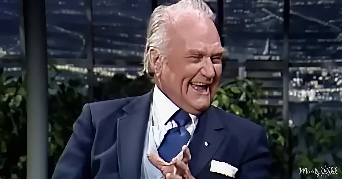 A man in a suit and tie is laughing heartily, holding his chest, with a cityscape and greenery in the background.