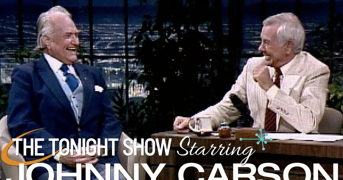 Two men in suits are seated, smiling, in a talk show setting with a cityscape background.