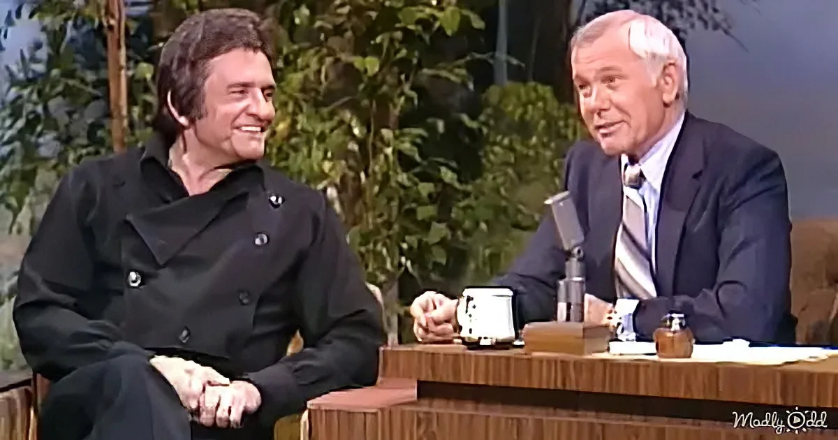 Johnny Cash in a black coat sits on a talk show set, smiling at the host.