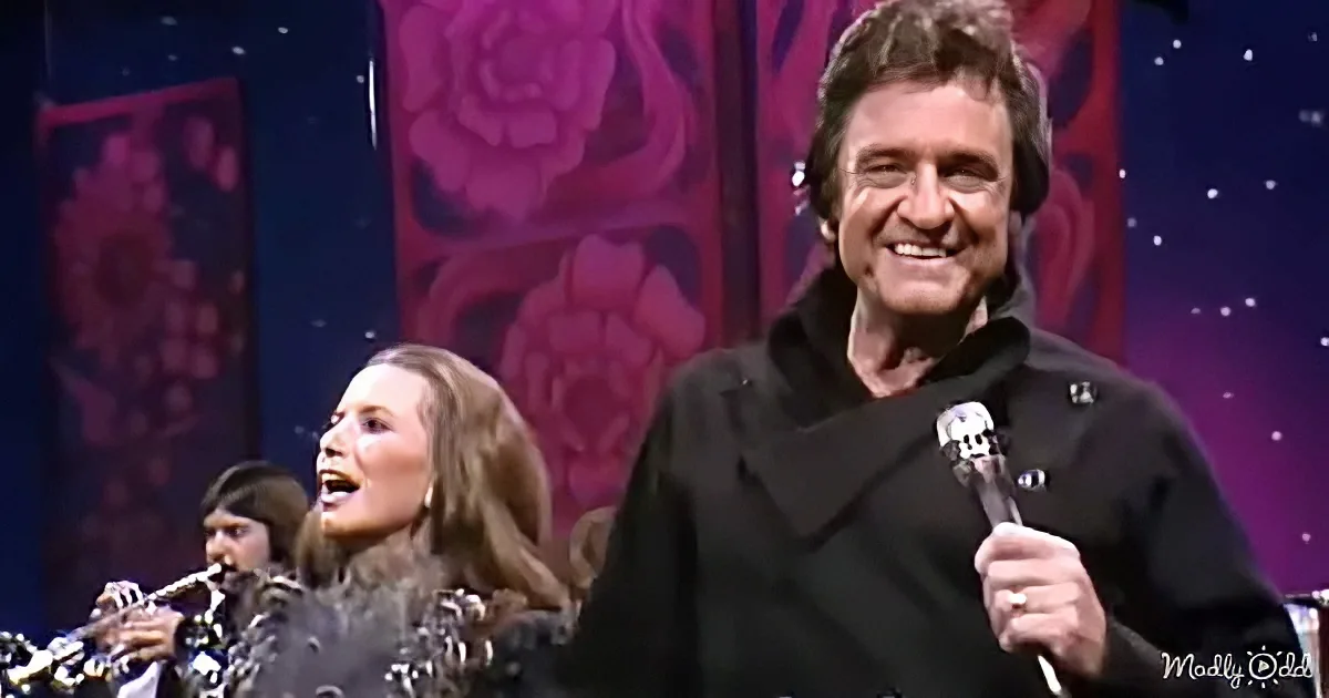 Johnny Cash holds a microphone, smiling, with a woman singing and a band in the background.