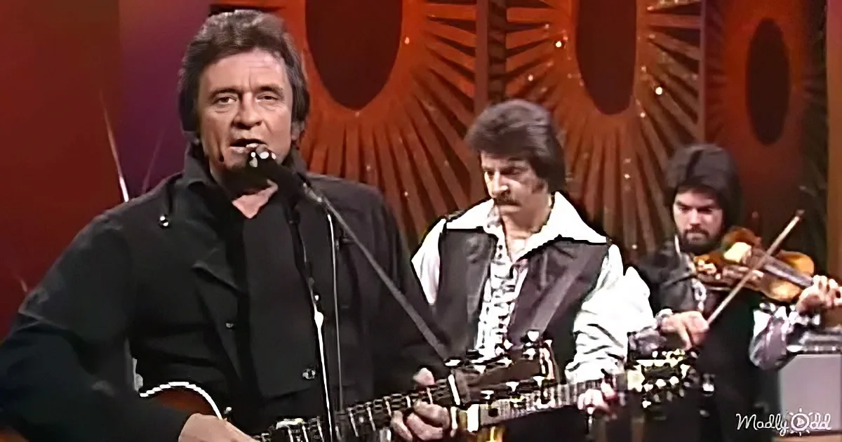 Johnny Cash plays guitar on stage with a band, wearing a black jacket, singing into a microphone.