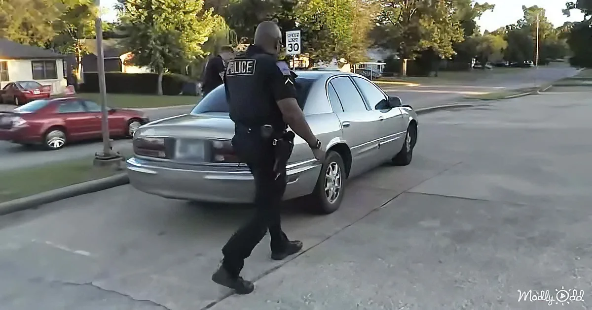 A police officer walks alongside a silver sedan on a suburban street.