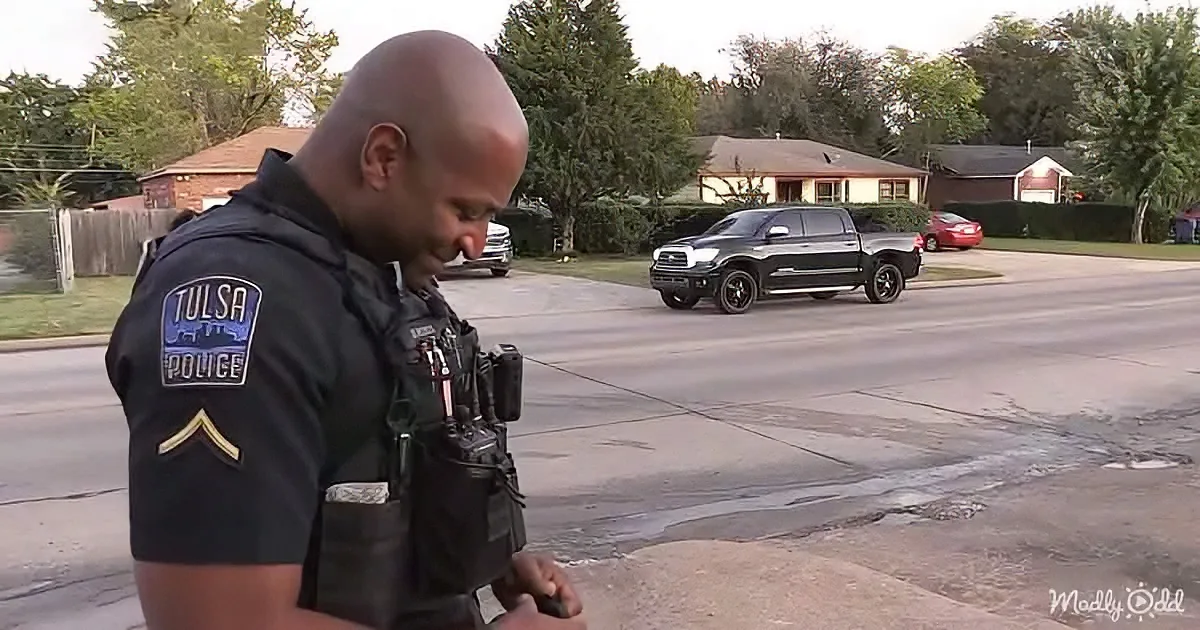 A Tulsa Police officer stands on a street corner, looking down, with a black pickup truck and houses in the background.