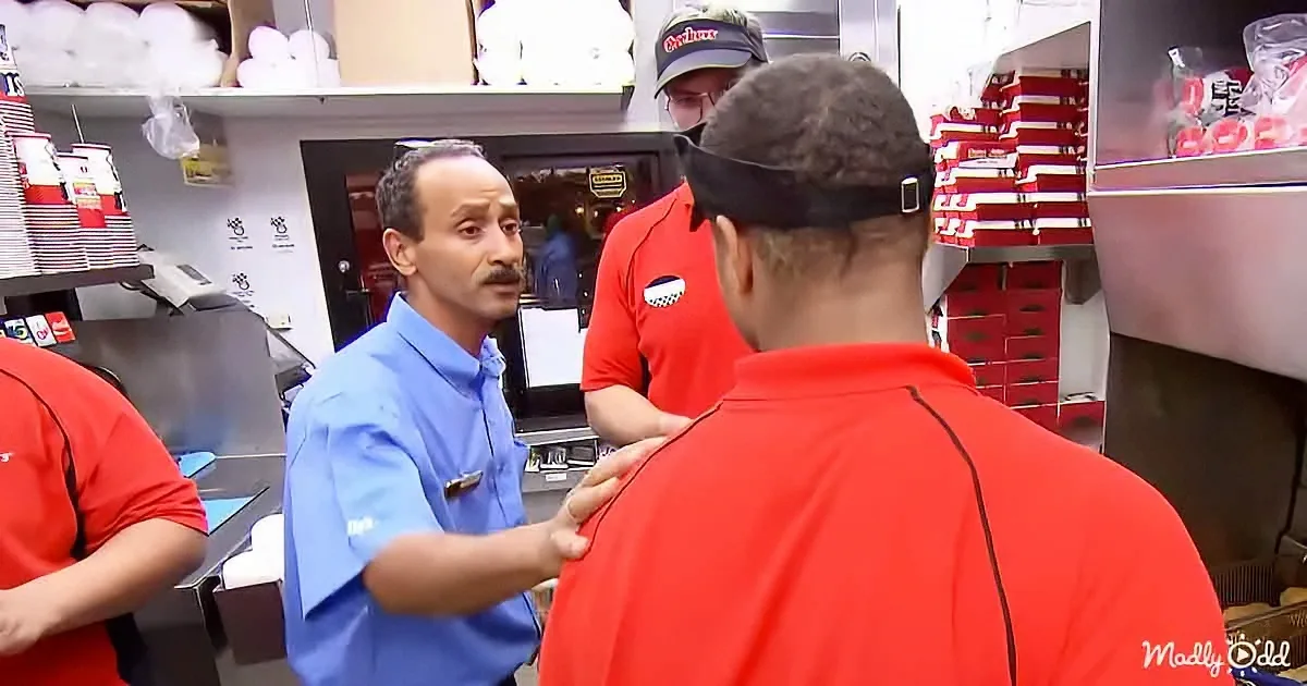 A man in a blue shirt is talking to a group of people in red shirts in a kitchen setting.