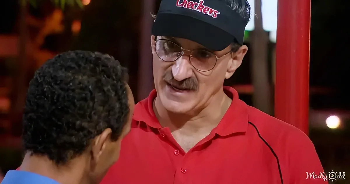 A man in a red Checkers uniform and cap is talking to another person with curly hair.