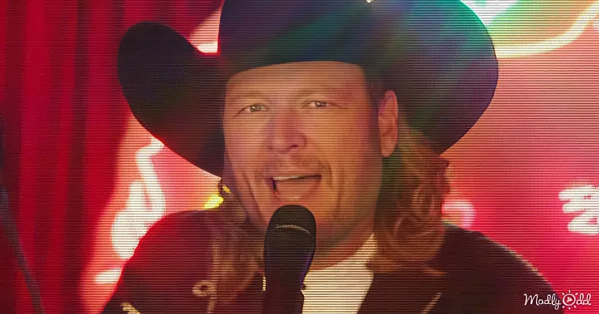A man in a cowboy hat and long hair sings into a microphone against a red curtain backdrop.