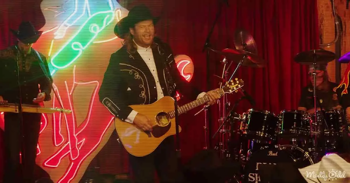 A man in a cowboy hat plays guitar on stage with a colorful neon backdrop and band behind him.