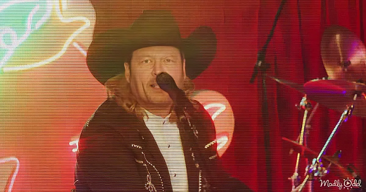 A man in a cowboy hat sings into a microphone on stage with a red backdrop and musical instruments.