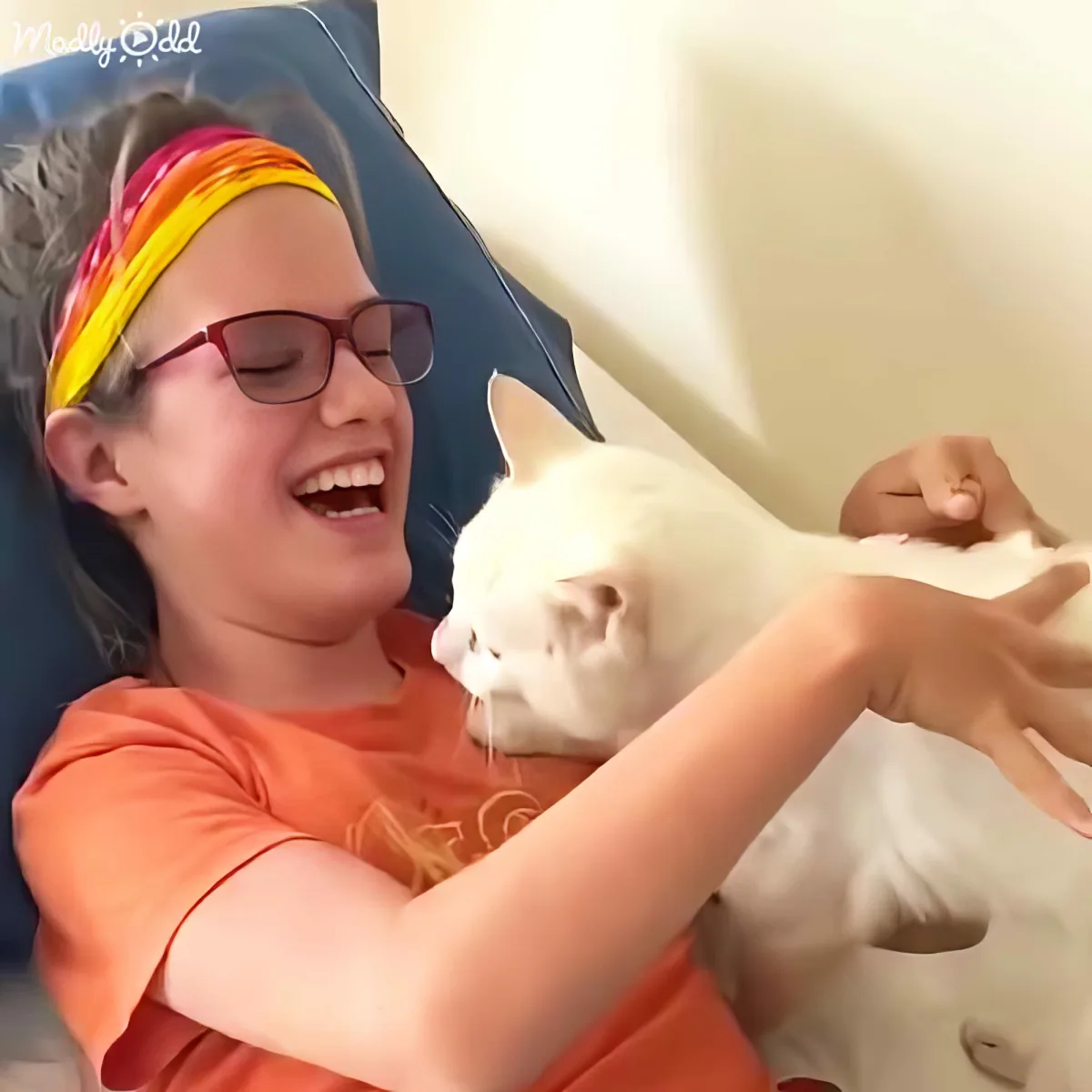 A person in an orange shirt and sunglasses is playfully interacting with a white cat, both smiling.