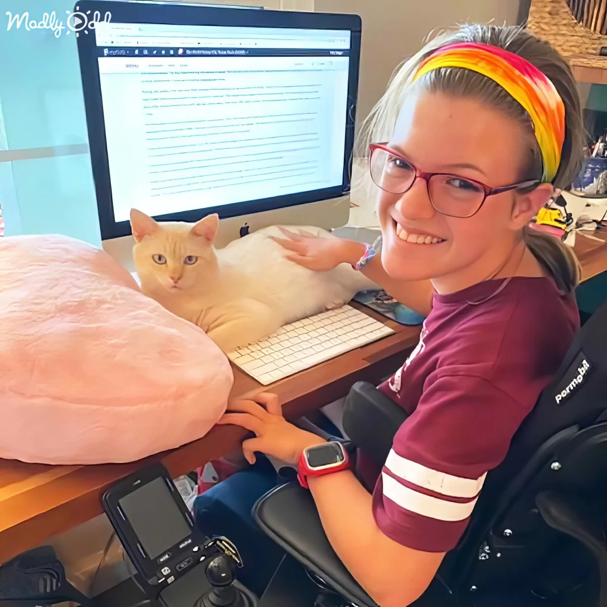 A smiling person in a wheelchair uses a computer with a cat lounging on the keyboard.