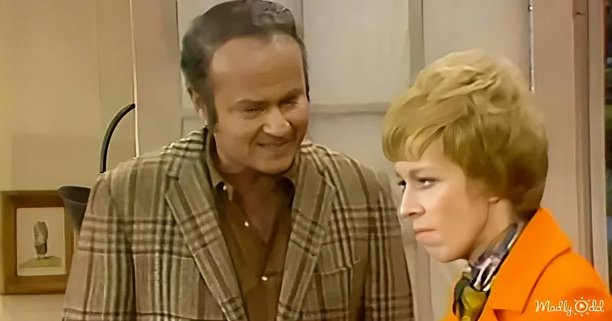 A man in a plaid blazer smiles at a woman in an orange jacket in a kitchen setting.