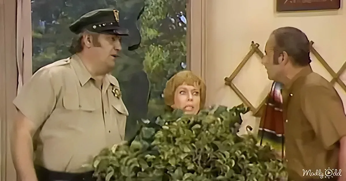 A man in a sheriff's uniform stands next to another man in a brown shirt, both looking at a surprised woman peeking from...