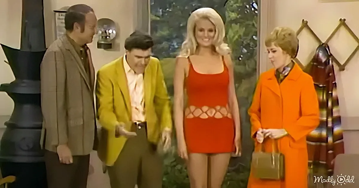 A woman in a red dress stands with three others in a room, one in a yellow suit holding a small object.