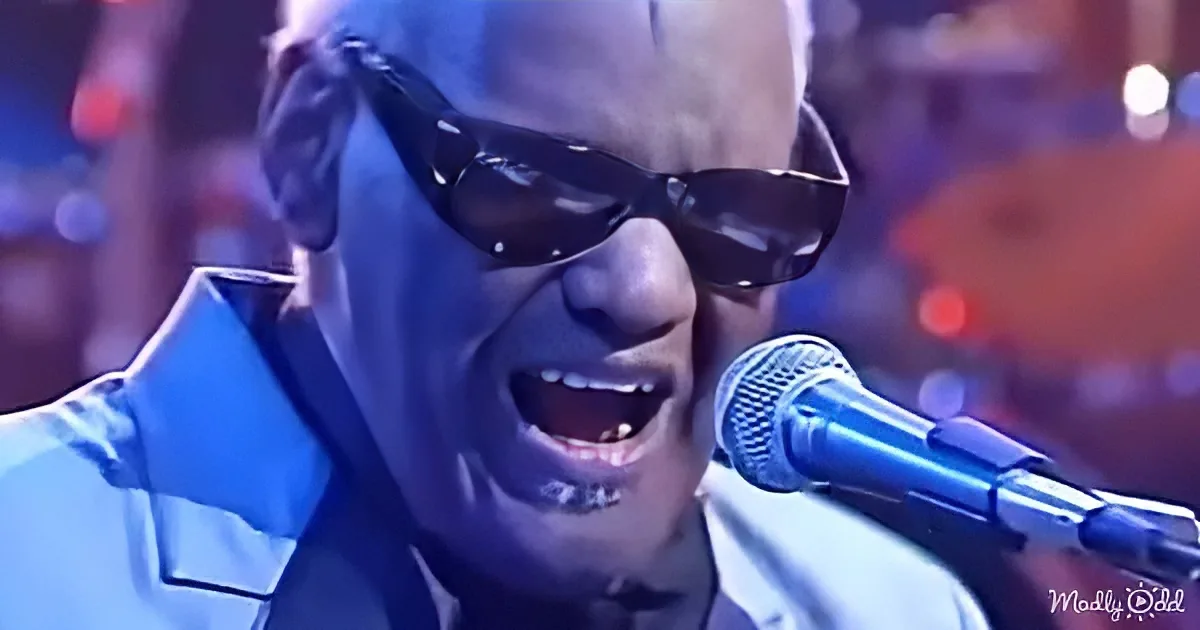 A man wearing sunglasses sings into a microphone, illuminated by blue stage lighting.