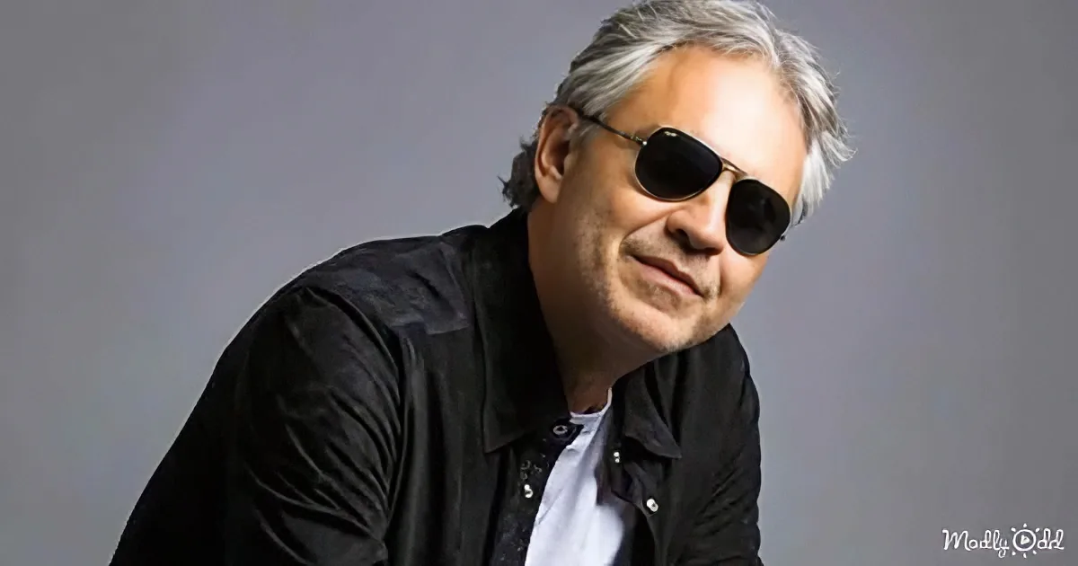 A man with gray hair and sunglasses, wearing a black jacket over a white shirt, is seated against a gray background.
