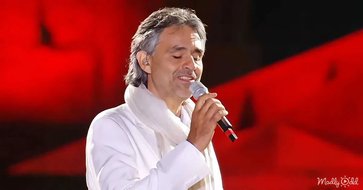 A man in white attire sings into a microphone on stage with red lighting.