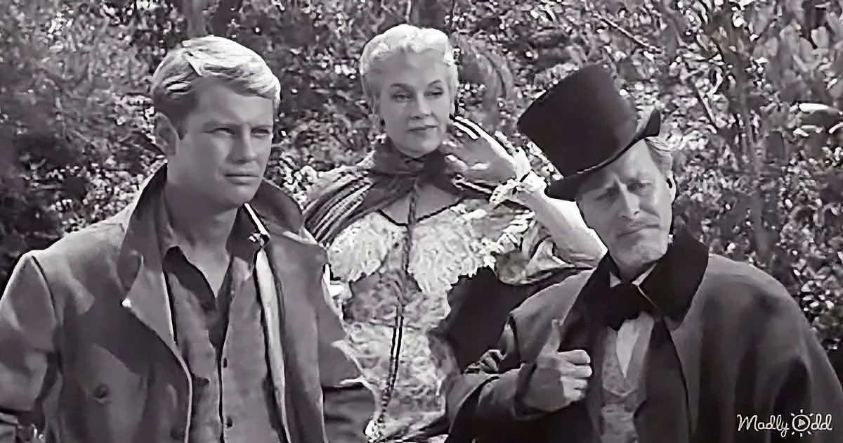 A man in a top hat and coat adjusts his hat while another man in a jacket looks on, with a woman in period attire in the...