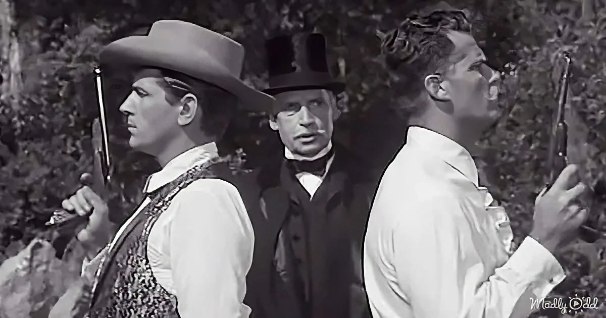Three men in Western attire, one holding a gun, stand in a forested area, seemingly in a tense moment.