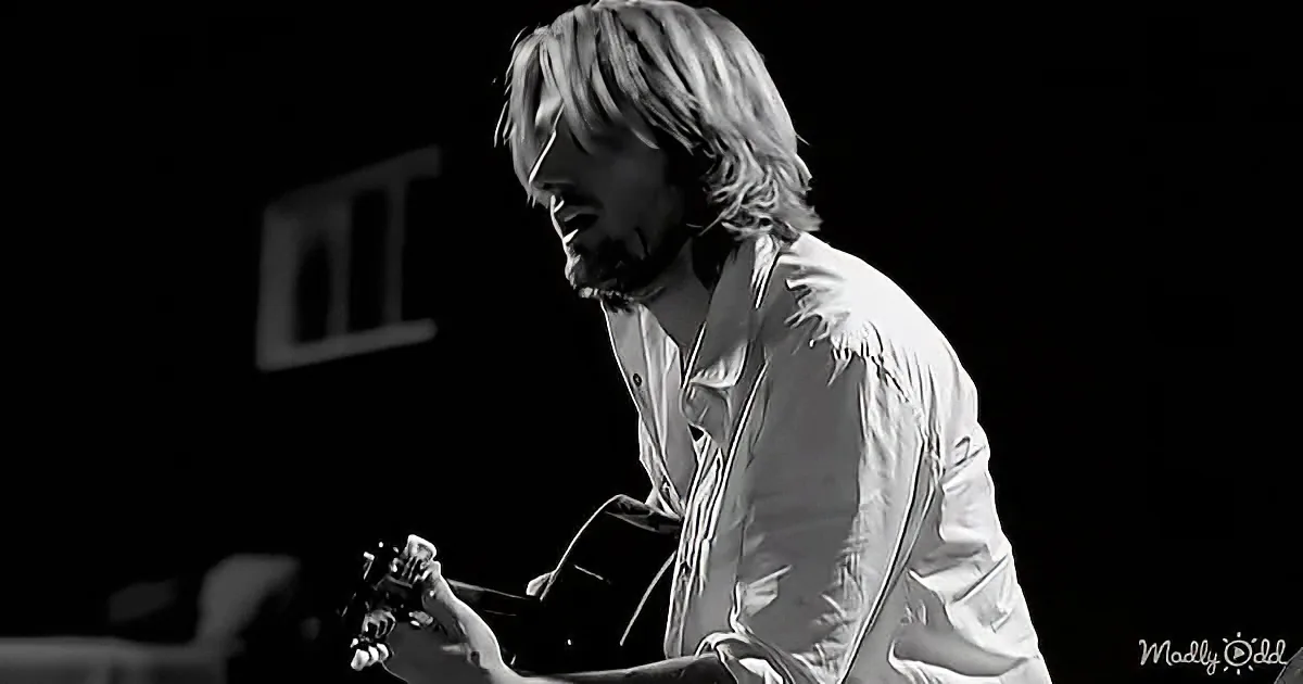 A man with long hair and a beard plays guitar, wearing a white shirt, in a black-and-white image.