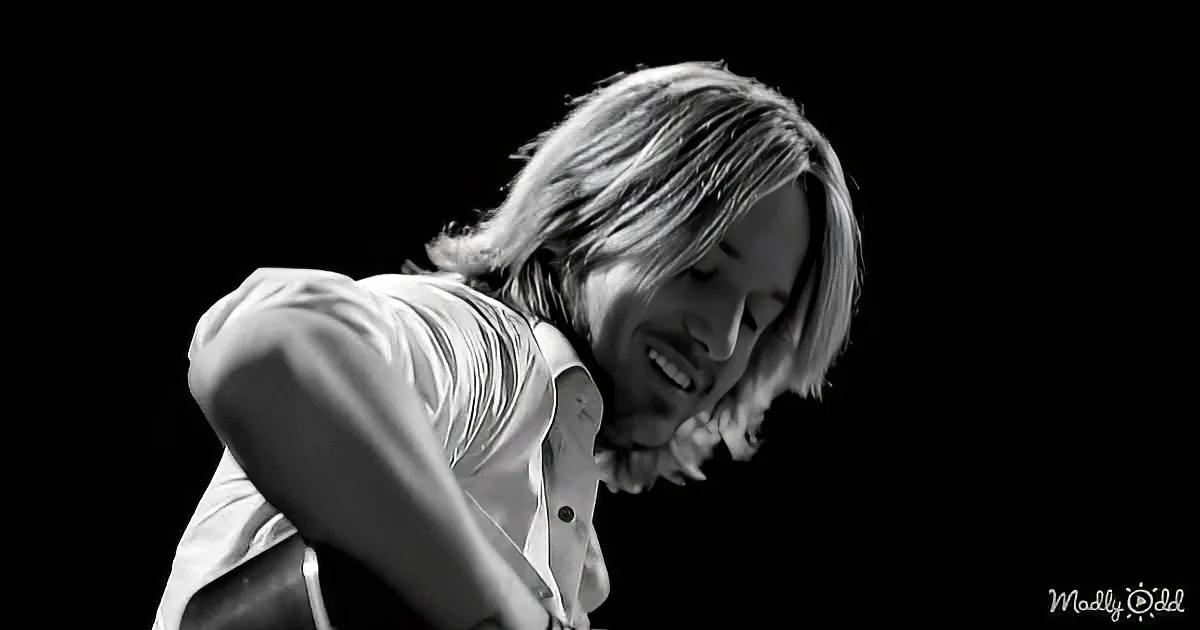 A person with long hair, wearing a white shirt, is playing a guitar against a dark background.