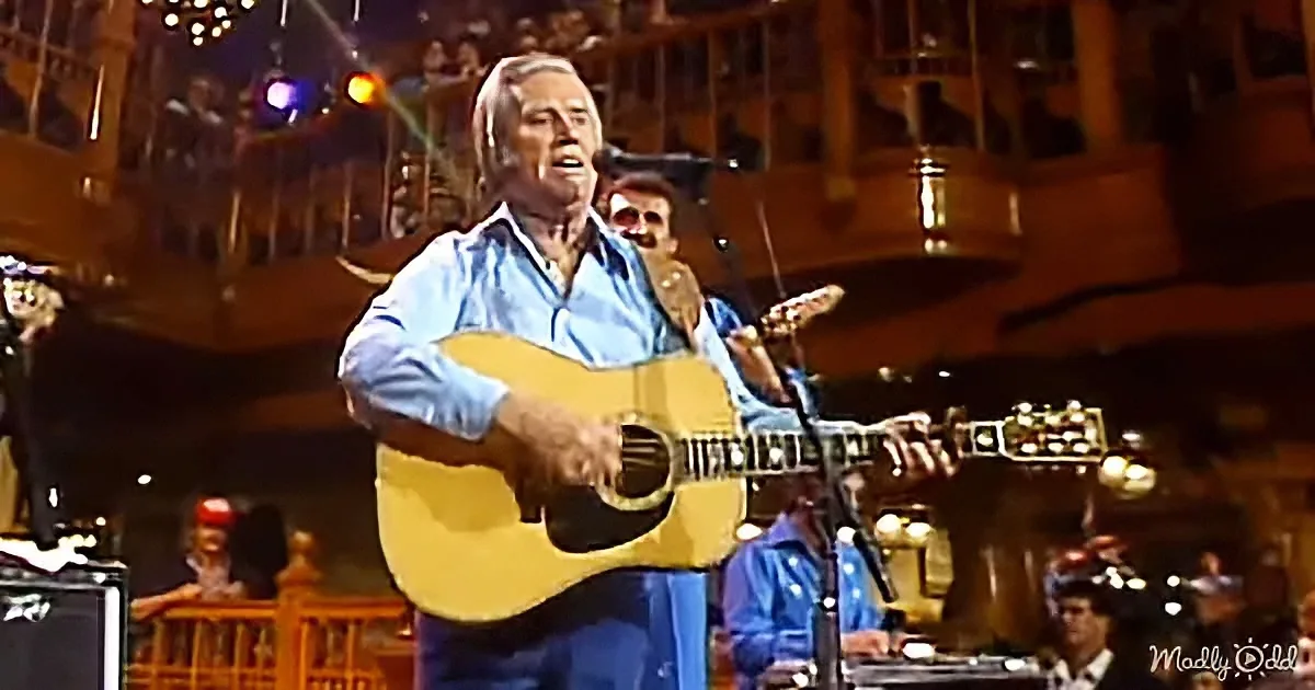 A man in a blue shirt plays an acoustic guitar on stage, singing into a microphone.