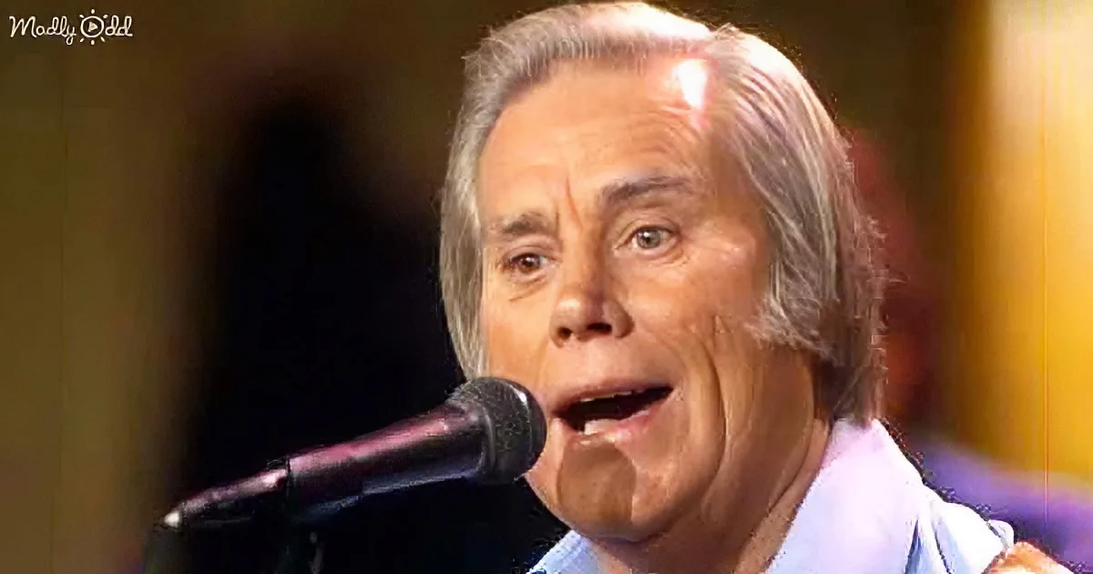 A man with gray hair sings into a microphone, wearing a light blue shirt, in a dimly lit setting.