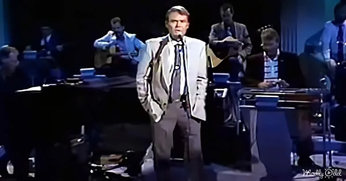 A man in a suit sings on stage with a band, including a guitarist and a drummer.