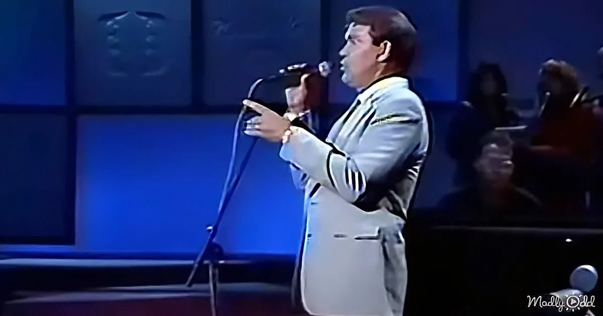 A man in a white suit sings into a microphone on stage.