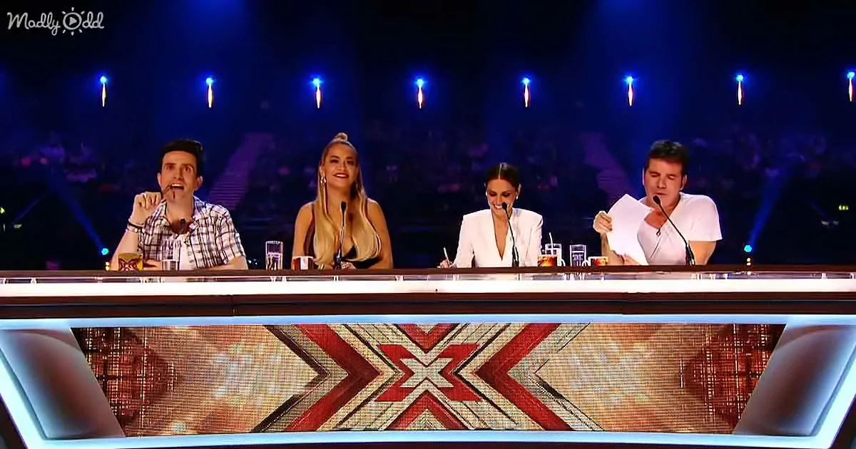 Four judges sit at a panel with microphones, one holding papers, in a dimly lit studio setting.