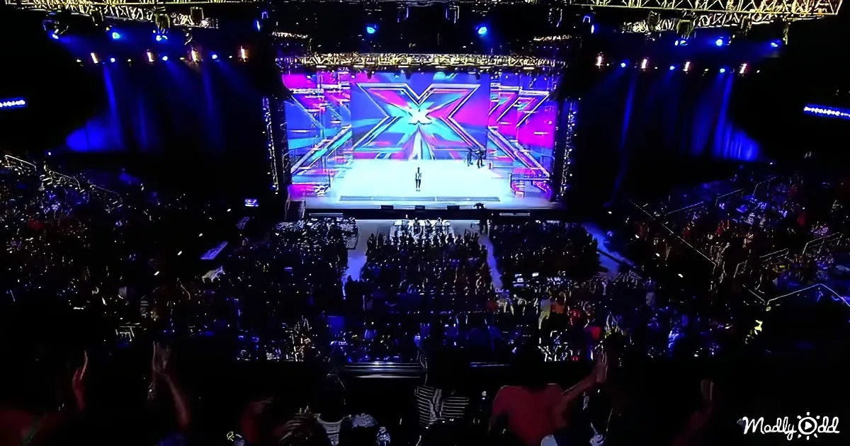 A performer stands on a brightly lit stage with a large "X" logo, surrounded by an excited audience in a dimly lit arena.