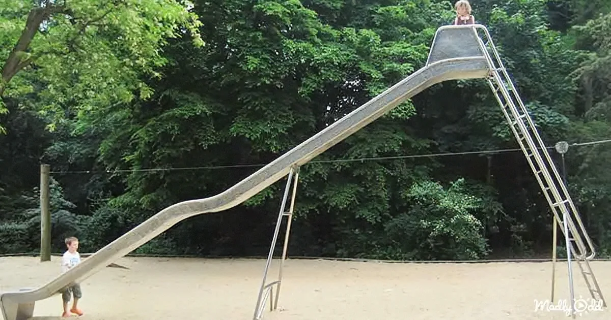 A child stands at the top of a large, curved slide in a park, preparing to slide down.