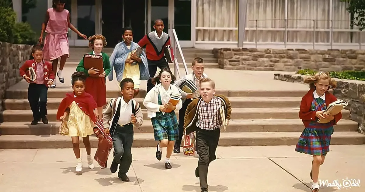 Children in school uniforms run down steps, holding books and bags, outside a stone building.