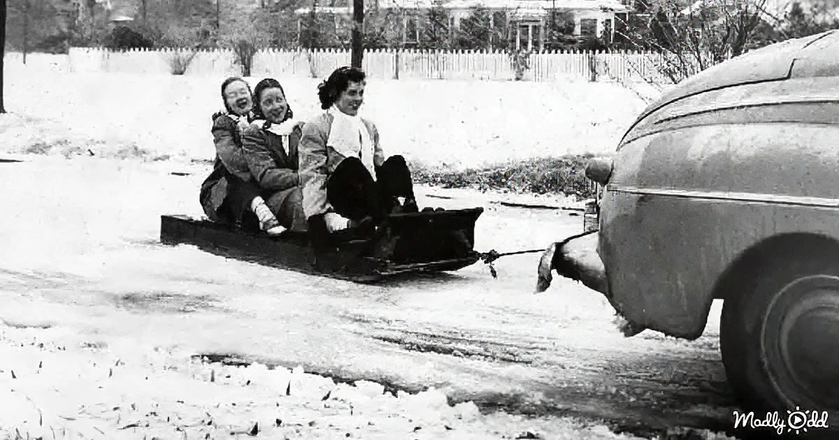 Three people are joyfully riding a sled being pulled by a car on a snowy street.