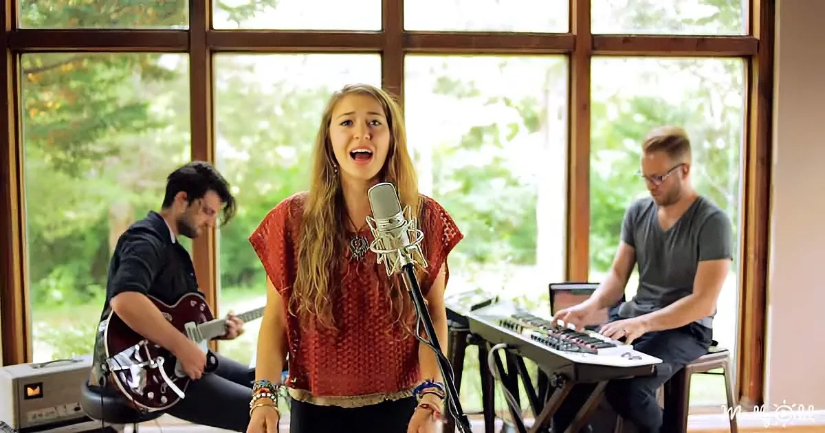 A woman sings into a microphone, accompanied by a guitarist and keyboardist in a room with large windows.