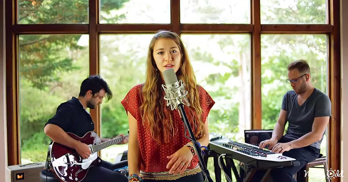 A woman sings into a microphone, accompanied by a guitarist and keyboardist in a room with large windows.