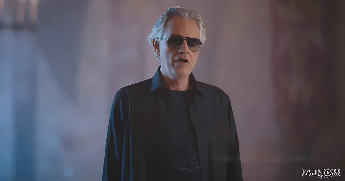 A man with gray hair and sunglasses stands in a dimly lit room, wearing a black shirt.