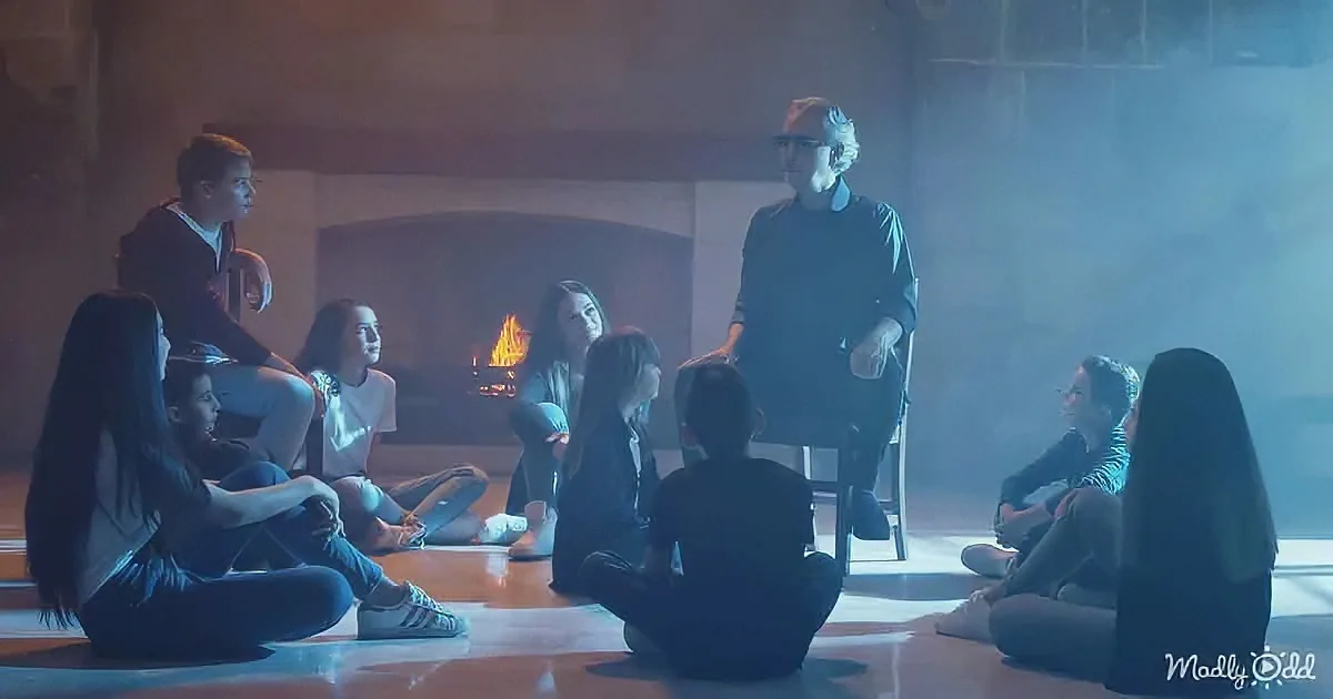A man stands in a dimly lit room, surrounded by a group of seated individuals, with a fire in the background.