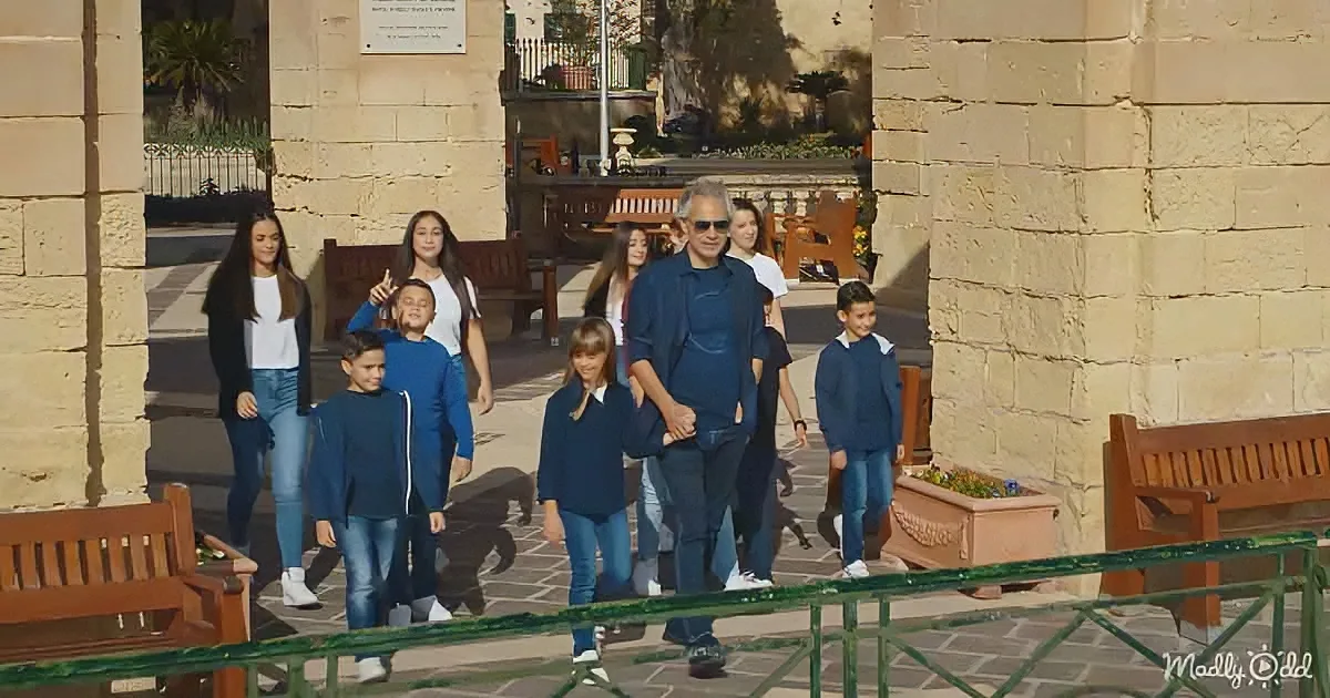A man walks with a group of children and adults in a sunlit courtyard, surrounded by stone walls and benches.